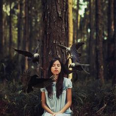 a woman sitting in the middle of a forest with birds on her shoulders and hands behind her head