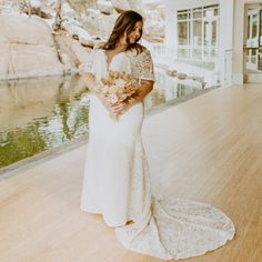 a woman in a white dress standing on a wooden floor next to a pond holding flowers