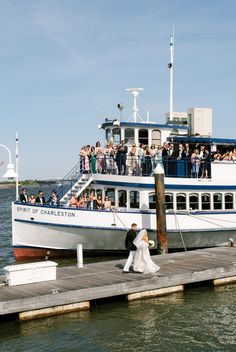 a bride and groom walking on the dock next to a boat with people in it