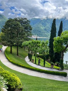 people walking down a path in the middle of a lush green park next to water