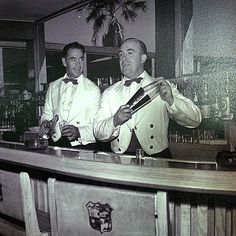 two men in tuxedos stand at the bar with their arms crossed and one man is holding a pair of scissors