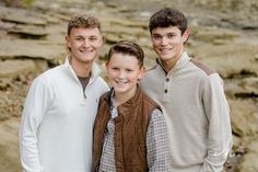 three young men standing next to each other in front of some rocks and water with one man smiling at the camera