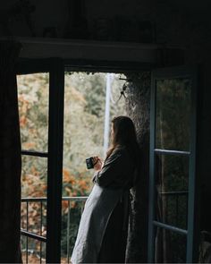 a woman standing in front of an open door holding a cup and looking out the window