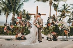 a bride and groom kissing in front of a cross surrounded by palm trees on the beach