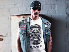 a man wearing a biker vest and bandana standing in front of a brick wall