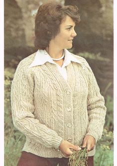 a woman standing in the grass wearing a white sweater and holding a plant with her hands