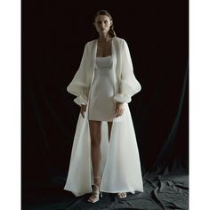 a woman standing in front of a black backdrop wearing a white dress and jacket with long sleeves