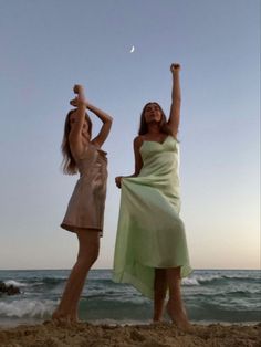 two women standing on the beach with their arms in the air and one holding her leg up