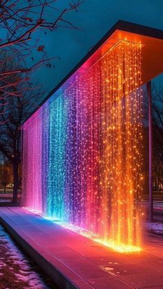 an illuminated water fountain in the middle of a park with trees and snow around it