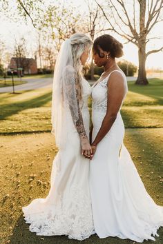 two women in wedding gowns standing next to each other on grass with trees behind them