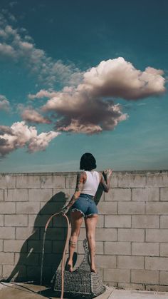 a woman standing on top of a cement wall next to a brick wall with clouds in the sky