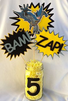 a mason jar filled with yellow popcorn and topped with batman cutout toppers on a white table cloth