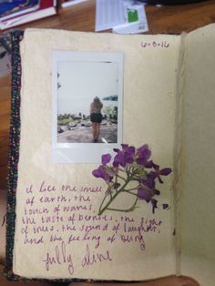 an open book with writing on it and a purple flower in the middle, sitting on a table