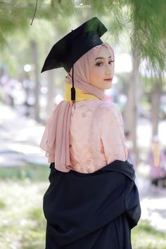 a woman wearing a black graduation gown and a yellow scarf on her head is standing under a tree