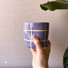 a hand holding a purple coffee cup with white lines on the outside, and a green plant behind it