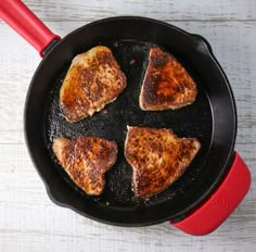 four pieces of chicken cooking in a skillet on a white wooden table with a red spatula