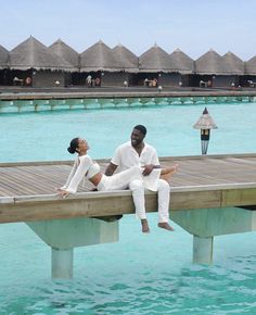 a man and woman sitting on a dock in front of water with thatched huts