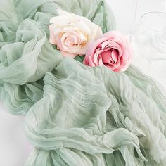 two pink and white flowers sitting on top of a green scarf next to wine glasses