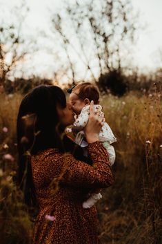 a woman holding a baby in her arms and kissing it's face while standing in tall grass