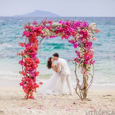 a bride and groom kissing on the beach in front of an archway with pink flowers