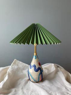 a green and blue lamp sitting on top of a white cloth covered tablecloth next to a gray wall