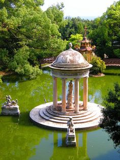 a gazebo in the middle of a green pond with statues on each side and trees around it