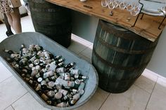 This shows a top down shot of a bar area display at a barn wedding. This focuses on a barrel bar, which are two large barrels with a wooden board over it. On the board are some glasses and drink dispensers. In front of the barrel is a large galvanised bath with beers in it. Prop Hire