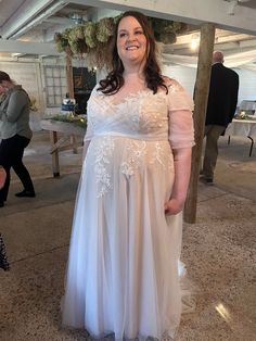 a woman wearing a white dress standing in a room with people walking around and looking at the camera