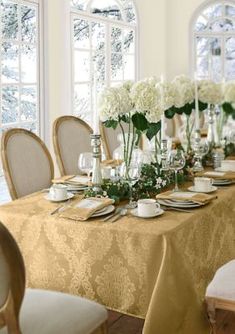 a long table with white flowers in vases and plates on it is surrounded by windows