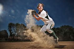 a female baseball player throwing a ball on a dirt field with trees in the background