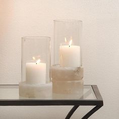 two white candles sitting in glass vases on top of a metal table next to a wall