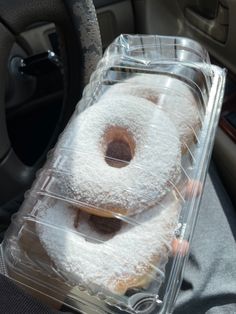 two donuts covered in powdered sugar sit in a plastic container on the dashboard of a car