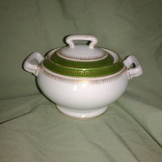 a white and green covered pot with handles on a tableclothed surface in front of a sheet