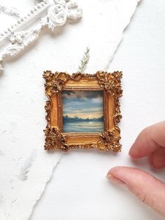 a hand holding a miniature painting in front of a white wall with an ornate gold frame