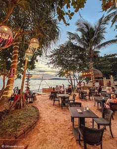 people are sitting at tables on the beach near the water and palm trees in front of them