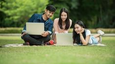 three young people sitting on the grass with laptops in their hands and looking at each other