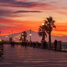 people are walking on the sidewalk at sunset with palm trees in the foreground and mountains in the background