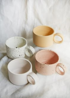 three different colored mugs sitting on top of a white bed sheet with black speckles