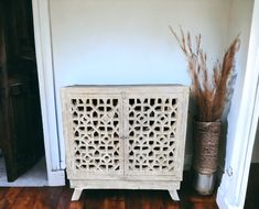 a white cabinet sitting on top of a wooden floor next to a vase with dried grass