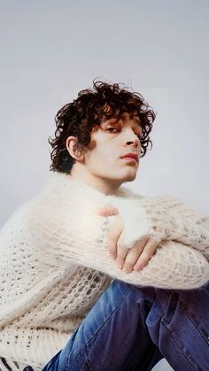 a man with curly hair sitting on the ground wearing a white sweater and blue jeans