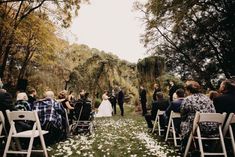 an outdoor wedding ceremony with white flowers on the ground and people standing in chairs watching