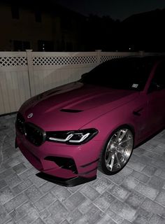 a pink car parked in front of a white fence at night with its lights on