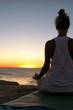 a woman sitting on top of a yoga mat in front of the ocean at sunset