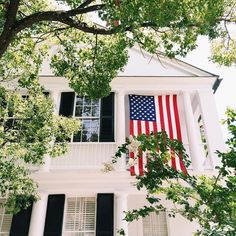 an american flag hanging from the side of a white house with black shutters and green trees