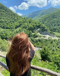 a woman standing on top of a wooden fence next to a lush green forest covered hillside