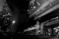 black and white photograph of city at night with light streaks on the buildings in the foreground