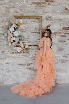 a woman in an orange dress standing next to a brick wall with flowers on it