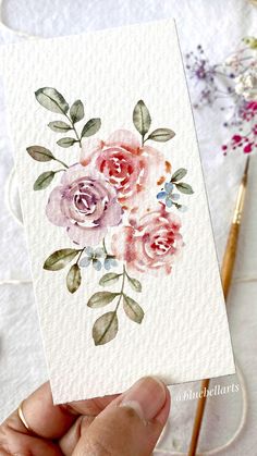 a hand holding up a card with watercolor flowers on it and a pen next to it