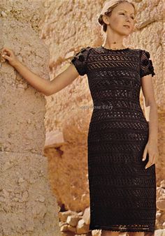 a woman standing next to a stone wall wearing a black crocheted dress with short sleeves