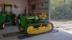 two green tractors parked in a garage next to each other with trees behind them on the side of the building